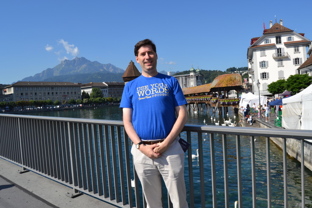 Photograph of Joshua Paul Cane wearing a James River Writers tee shirt posing before the Kapellbrücke in Lucerne, Switzerland.